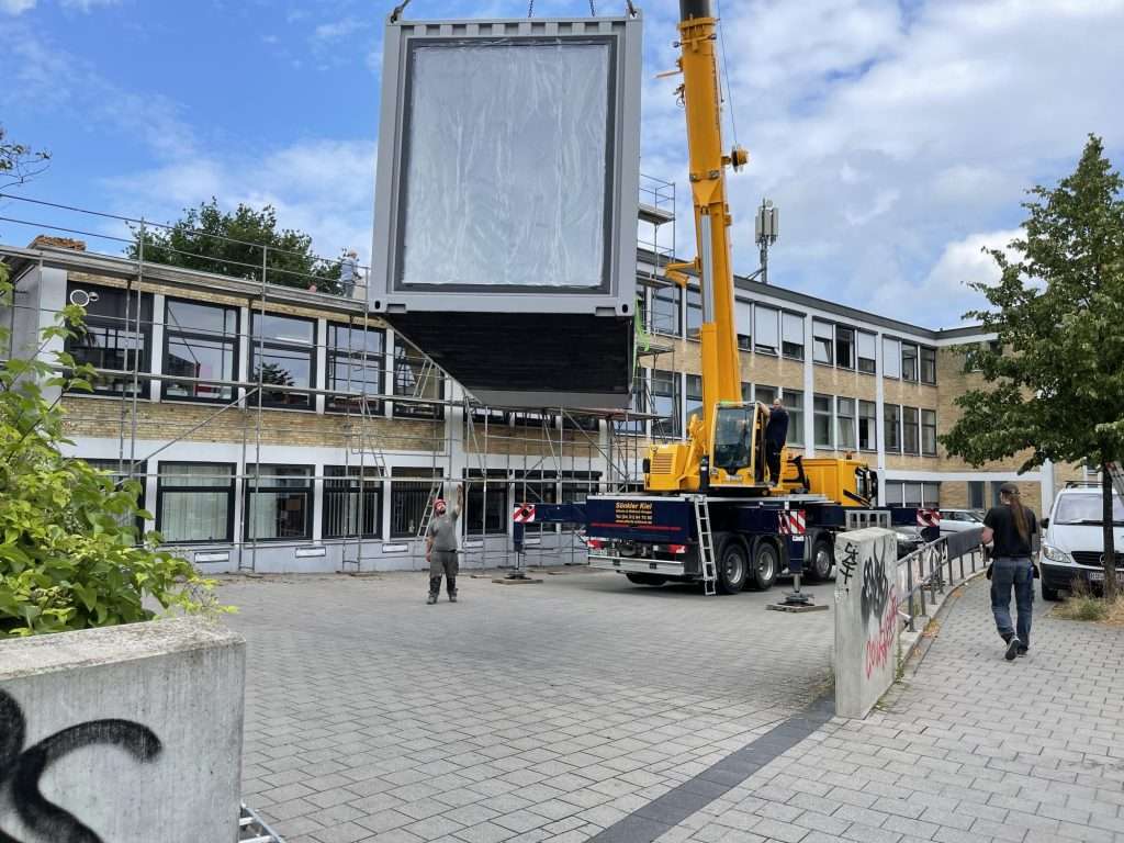 Container module hanging from a crane in front of a building