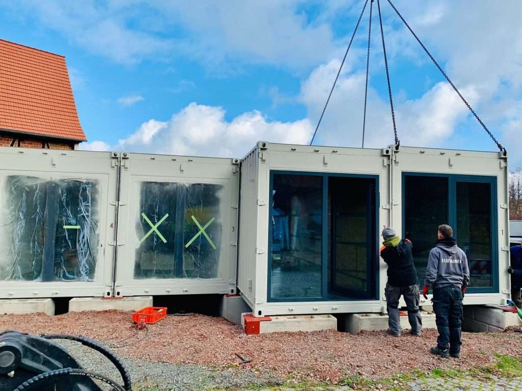 Container modules with glass doors on foundations while a further module is lifted into place