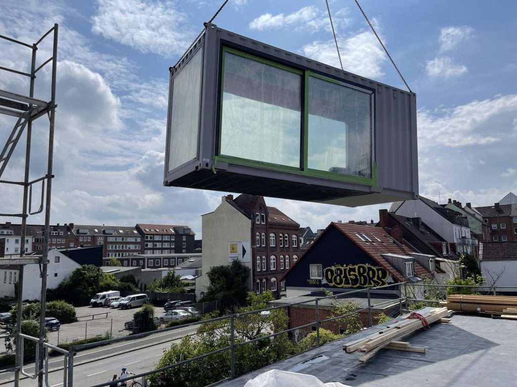 Crane lifting a container module with sliding windows above the roofs of a city