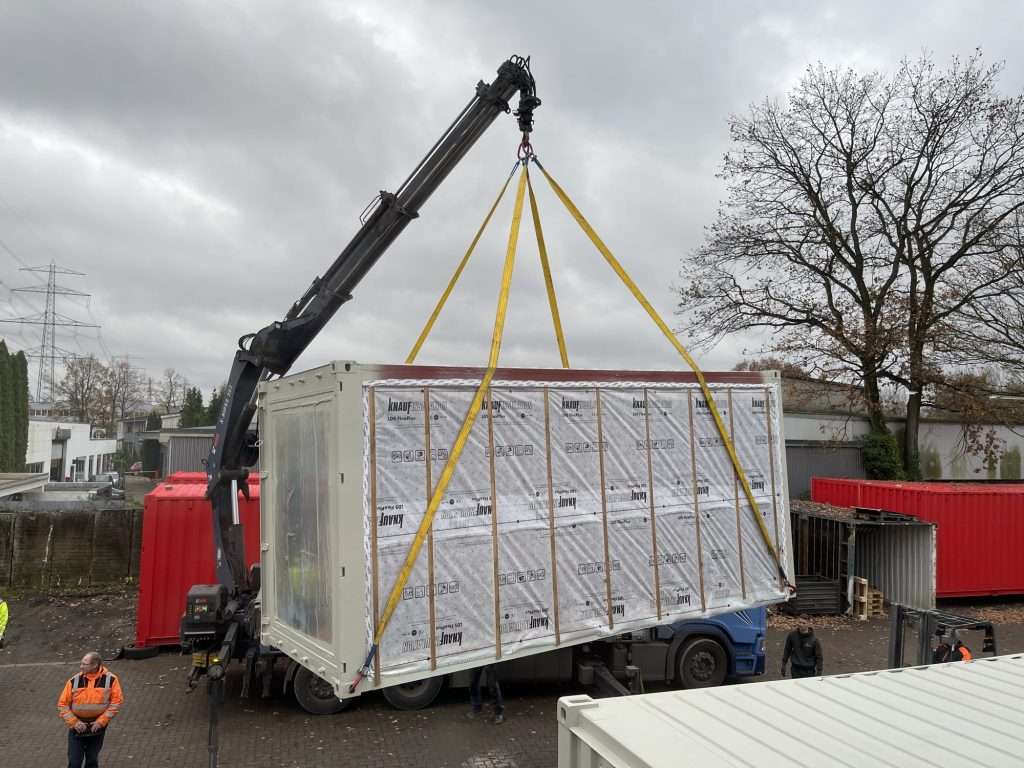 Loader crane lifting a container module wrapped in insulation board and film off a truck