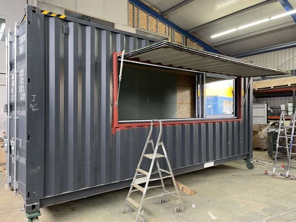 Dark grey shipping container with a window opening and a steel hatch cover in a workshop
